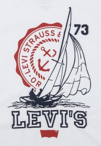 Camiseta blanca con un gráfico de yate en rojo y azul marino, logo circular de Levi's y texto bordado "73" en el lado derecho.