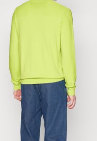 Man i limegrön långärmad tröja och löst sittande blåjeans, sedd bakifrån mot en enfärgad bakgrund.