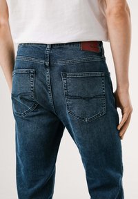 Los vaqueros de denim azul presentan una cintura de altura media, costuras en contraste y dos bolsillos traseros. Se puede ver una etiqueta de la marca en la cinturilla.