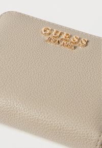Jasnobeżowy portfel z wegańskiej skóry o fakturowanym wykończeniu, z wytłoczonym złotym logo "GUESS EST. 1981" oraz zaokrąglonymi krawędziami.