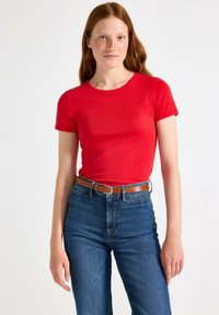 Jonge vrouw met lang rood haar die een rode top met korte mouwen, blauwe spijkerbroek en een bruine riem draagt, staande tegen een effen witte achtergrond.