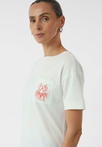 T-shirt blanc en coton doux, avec une poche ornée d'un design de homard rouge brodé. Manches courtes et col rond.