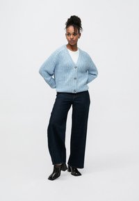 Cardigã de malha azul claro com decote em V e três botões grandes, combinado com jeans escuros de perna larga e botas de salto pretas brilhantes até ao tornozelo.