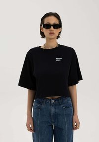 Zwarte cropped t-shirt met korte mouwen, voorzien van een klein wit logo, gedragen met hooghangende blauwe jeans met contrasterende stiksels.