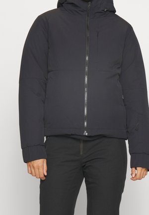 Homme portant une veste noire matelassée à capuche avec une fermeture éclair frontale et une poche poitrine zippée, associée à un pantalon noir.