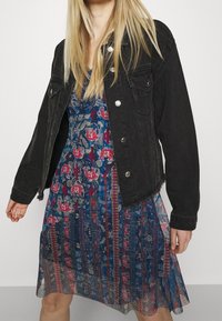 Svart denimjacka med silverknappar, kombinerad med en blå mönstrad klänning som har röda elefanter och blommönster. Långt hår synligt.