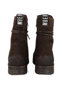 Brown suede ankle boots featuring a round toe, stacked heel, and a decorative black and white band at the back. Simple lace detail.