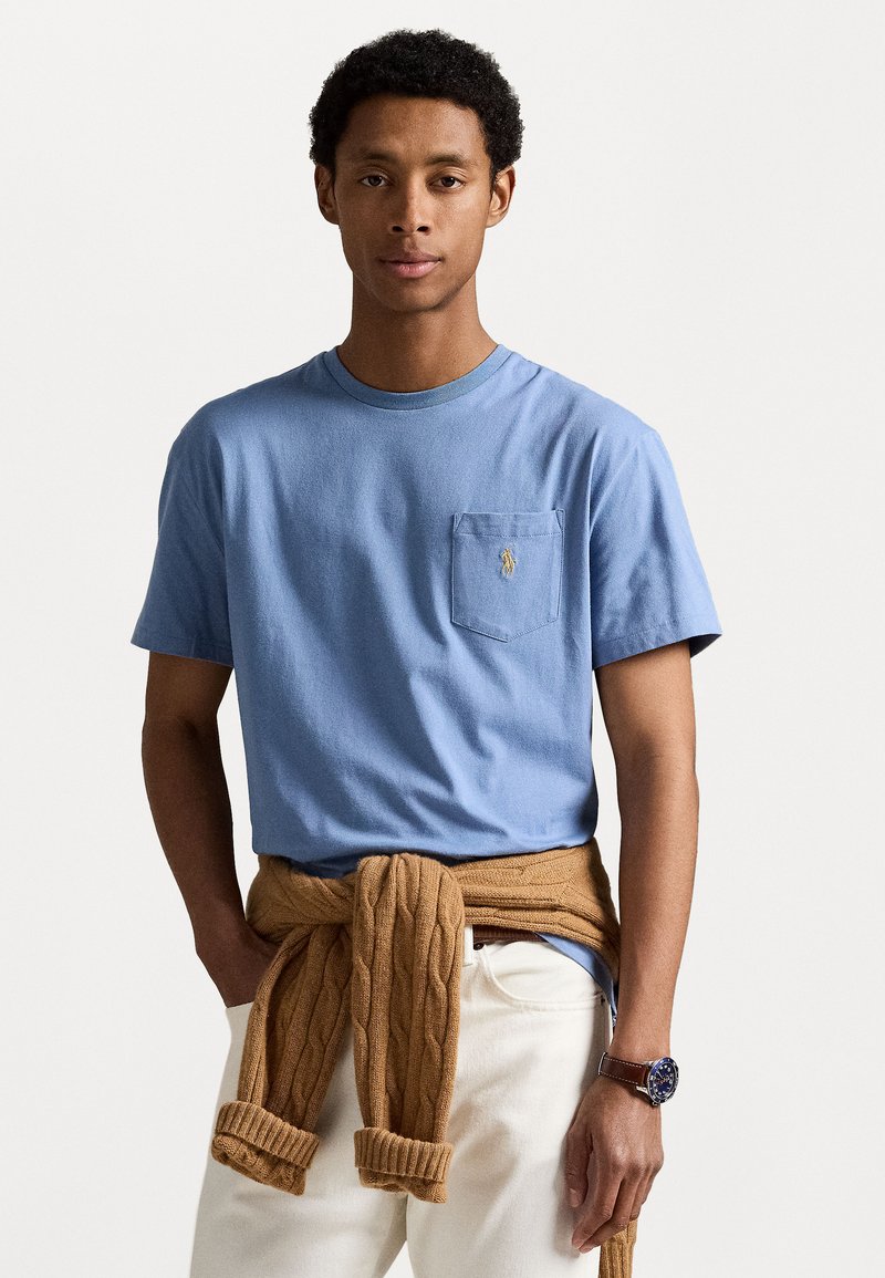 Tricou din bumbac de culoare albastru deschis, cu un buzunar pe piept și un logo brodat, asortat cu un pulover tricotat de culoare maronie legat la talie și pantaloni albi.