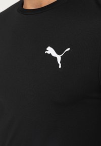 Camiseta deportiva negra hecha de tela suave, con un logotipo blanco de Puma en la parte superior izquierda. Cuello redondo, diseño ajustado.