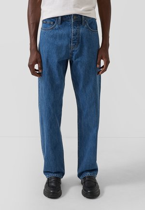 Man draagt losse blauw denim jeans en zwarte loafers tegen een effen lichte achtergrond.