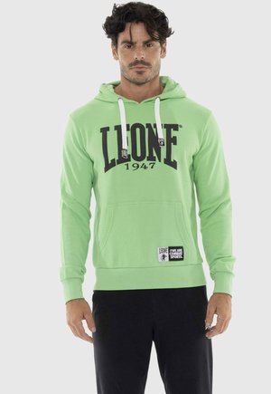 Uomo che indossa una felpa con cappuccio Leone 1947 verde chiaro con cordoncini bianchi e pantaloni neri, in piedi contro uno sfondo neutro.