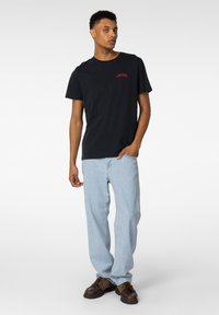 Svart bomulls T-skjorte med rød tekst, sammen med lyseblå denim jeans. Modell iført brune sko, står med den ene hånden i lommen.