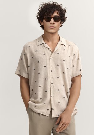 Mann mit lockigem Haar, der runde, dunkle Sonnenbrillen trägt und ein beigefarbenes Kurzarmhemd mit kleinen schwarzen Blumenmustern anhat.