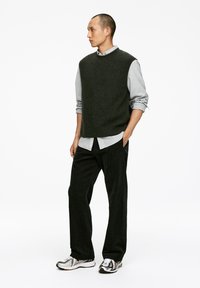 Dunkelgrüner Wollmischung ärmelloser Pullover über einem hellgrauen Hemd mit Kragen, kombiniert mit schwarzen Cordhosen und weißen Sneakern mit schwarzen Akzenten.