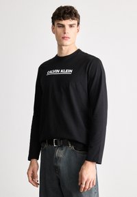 T-shirt de manga longa preta com o logótipo branco "CALVIN KLEIN". O tecido parece suave, com um corte clássico, combinado com jeans escuros.