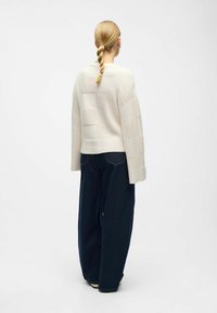 Suéter de punto color crema con un patrón de cuadrícula texturizado, combinado con unos pantalones vaqueros de corte relajado en azul oscuro. El suéter tiene mangas anchas.