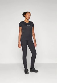 Noor naine seisab seljas fitted must spordivorm koos logodega, mustad jooksukingad ja neutraalne ilme, tasase heleda tausta ees.