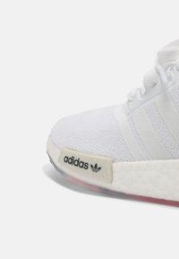 Vit löparskor med ett texturerat mesh-övre, tre vita sidostråk och en gummisula med en röd accent. Adidas-logotyp på hälen.