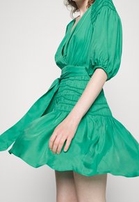 Vestido de seda verde con un escote en V profundo, mangas cortas abullonadas y cintura ajustada. Presenta una falda acampanada y un cinturón atado a juego.
