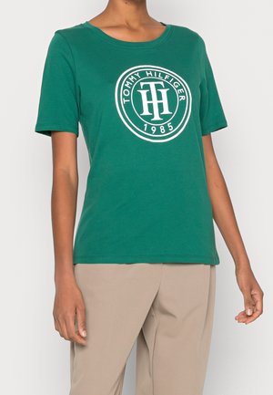 Osoba nosząca zieloną koszulkę z krótkim rękawem z białym logo Tommy Hilfiger 1985, w połączeniu z beżowymi spodniami na jednolitym tle.