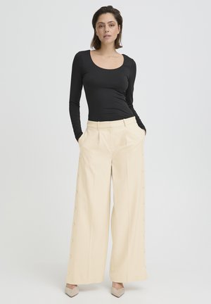 Donna in piedi con le mani in tasca, indossa una maglia nera aderente a maniche lunghe, pantaloni crema a vita alta e gamba larga, e scarpe beige con punta affilata.