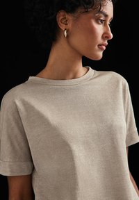 T-shirt beige a maniche corte con scollo rotondo, realizzato in un tessuto morbido e luccicante. Presenta risvolti sulle maniche e una vestibilità rilassata.