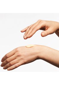Una crema di un beige chiaro è applicata su una mano con pelle leggermente texture. La mano è posizionata sopra il polso, con le dita leggermente curve.