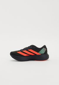 Baskets de sport noires avec des rayures rouges, tissu texturé, bout arrondi et design élégant. Les accents comprennent un panneau vert et une semelle rouge.