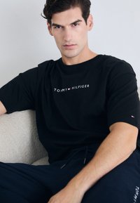 Camiseta de algodón negra de manga corta, con el texto del logo en blanco "TOMMY HILFIGER" estampado en el pecho y un pequeño detalle del logo en la manga.