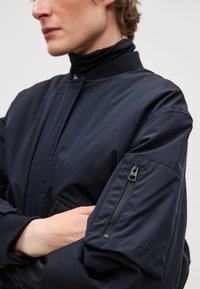 Tumšzils bumbvedēja jaka gludā audumā ar rāvējslēdzēja kabatu uz kreisās rokas, ribām apvilktiem aprocēm un augstu apkakles dizainu.