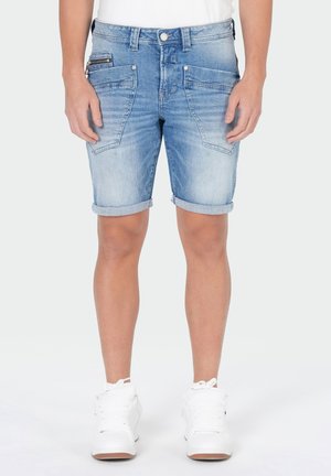 Homme portant un short en jean bleu clair retroussé avec de grandes poches avant et des baskets blanches, debout devant un fond uni.