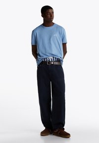 T-shirt di cotone azzurro chiaro con un piccolo logo sul petto, abbinata a pantaloni blu scuro e scarpe marroni. Design semplice e casual.