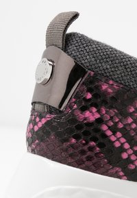 Primo piano di un tallone di sneaker con motivo in pitone rosa e nero, colletto in maglia grigia, linguetta metallica e suola bianca.
