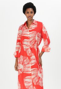 Rochie roșie cu butoane în față, cu modele florale supradimensionate în crem. Mâneci lungi, talie legată, țesătură ușoară și croială lejeră.