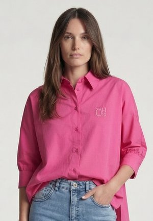 Frau mit langen braunen Haaren, die ein knallpinkes Hemd mit Knöpfen und hellblaue Jeans trägt, eine Hand in der Tasche, neutraler Gesichtsausdruck.