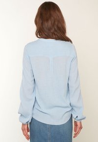 Lichtblauwe lange mouw shirt van textured stof, ronde hals en met knoopmanchetten. Beschikt over een transparante achterpaneel en een relaxed fit ontwerp.