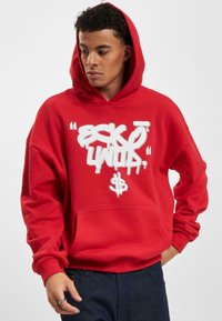 Felpa rossa con un grande logo bianco in stile graffiti sul davanti. Presenta una tasca a marsupio e un cappuccio con cordino. Tessuto morbido, vestibilità rilassata.