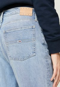 Jeans de denim azul claro con un parche de cuero etiquetado como "TOMMY JEANS", con un pequeño logo en el bolsillo trasero y un corte relajado.