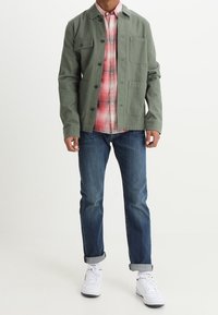 Kurtka koszulowa w oliwkowej zieleni z zapięciem na guziki i dwoma kieszeniami na piersiach, nałożona na czerwoną koszulę w kratę, zestawiona z niebieskimi dżinsami i białymi trampkami.