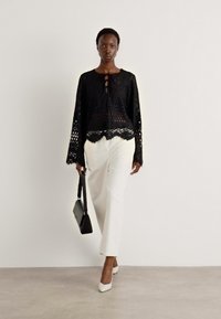 Svart blonde topp med flagrende ermer og scalloped kant, kombinert med hvite bukser og spisse hæler. Modellen bærer en svart håndveske.