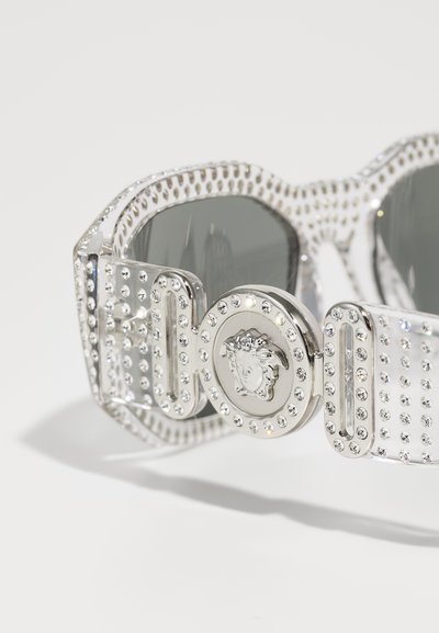 Silver sunglasses with a jeweled frame, featuring a gray lens and a circular embellishment with a lion's head design.