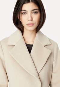 Beige Wollmischungsmantel mit Reverskragen, weich im Griff und tailliert geschnitten, über einem dunklen Oberteil getragen. Schlichtes, minimalistisches Design.