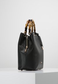 Černá kožená kabelka s texturovaným povrchem, zlatým zipem a hnědými rukojeťmi ve stylu bambusu, ležící na bílé podstavě na pozadí bez vzoru.