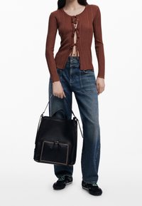 Žena v hnědé žebrované kardiganu s uvázáním, modrých džínech s vysokým pasem, černých teniskách, držící velkou černou koženou tašku.