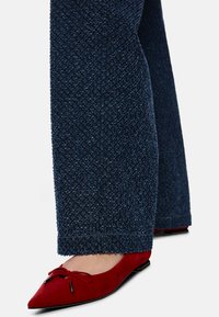 Marineblaue Hosen aus strukturiertem Stoff, kombiniert mit roten Ballettschuhen aus Wildleder, die eine Schleifenverzierung und schwarze Hardware-Details aufweisen.