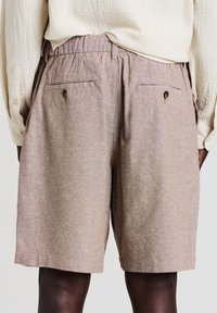 Eine Person trägt beige Leinenshorts mit elastischem Bund und zwei hinteren Knopf-Taschen, kombiniert mit einem cremefarbenen, strukturierten Langarmhemd.