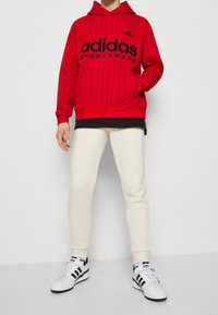 Sudadera con capucha roja con rayas verticales negras y gran logo de Adidas, combinada con pantalones de chándal blancos y zapatillas en blanco y negro.