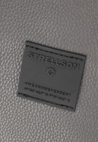 Zwart gestructureerd leren patch met het reliëflogo "STRELLSON", een hashtag en coördinaten, genaaid op een gladde grijze leren achtergrond.