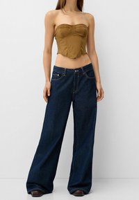 Donna che indossa un top corsetto marrone senza spalline e jeans larghi blu scuro con scarpe nere su uno sfondo neutro.