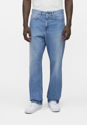 Μοντέλο μεσαίας ένδυσης φοράει ανοιζτό μπλε τζιν με ίσια μπότα και πέντε τσέπες, συνδυασμένο με λευκά αθλητικά παπούτσια και λευκό πουκάμισο που είναι μπαζωμένο μέσα.